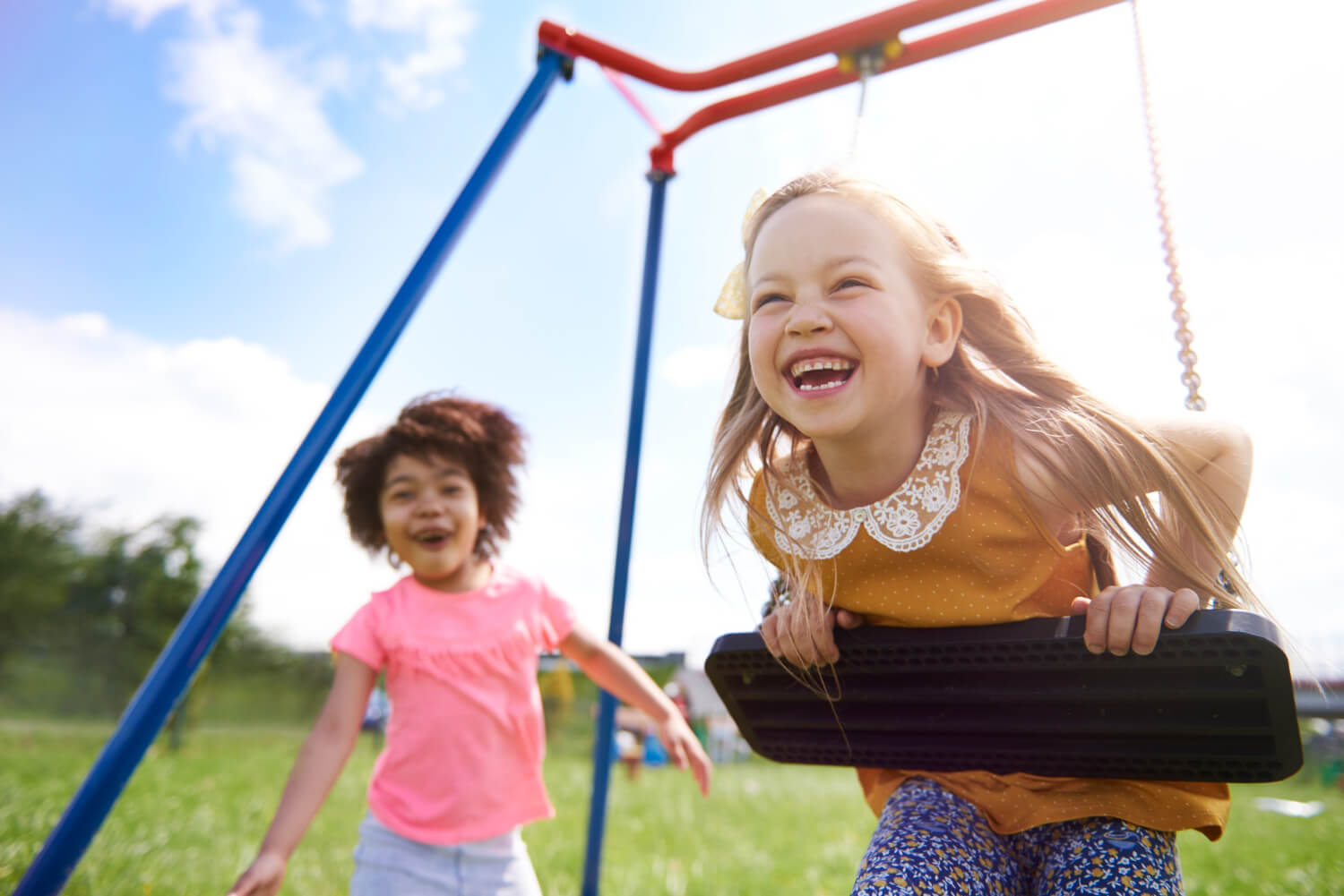 Menina loira sorrindo em um balanço de parque enquanto outra criança corre ao fundo. Ideal para prevenir trauma dental em Pato de Minas.